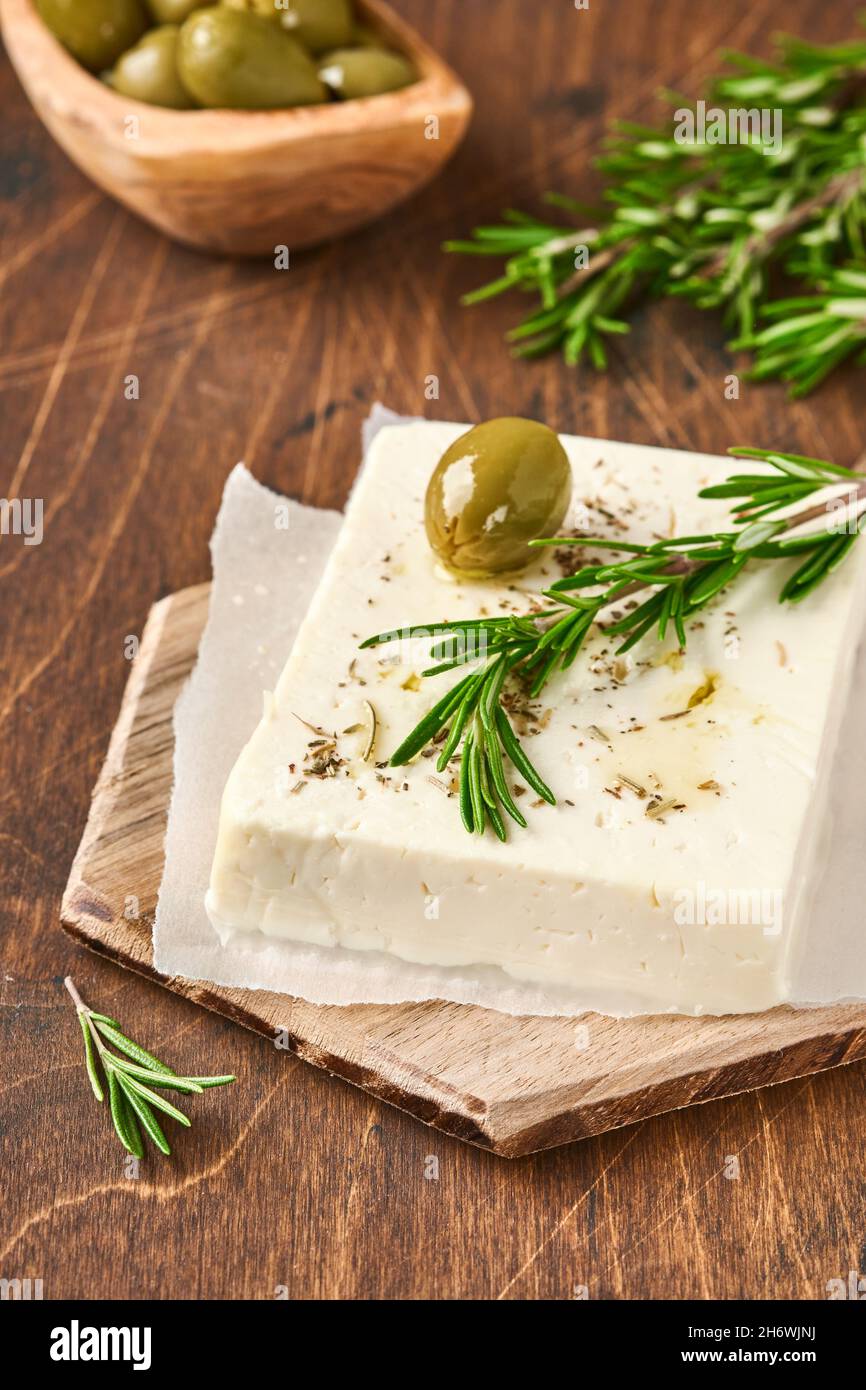 Cheese feta with rosemary, herbs, olives and olive oil on wooden cutting board on old wooden background. Traditional Greek homemade cheese. Selective Stock Photo