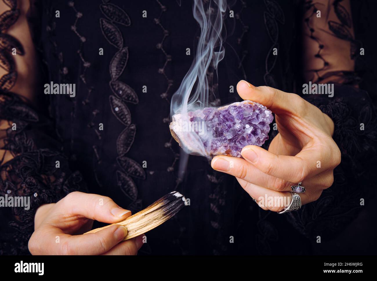 Close up view of woman in black lace dress, cleansing amethyst crystal cluster gemstone by smudging Palo Santo wood stick. Remove negative energy. Stock Photo