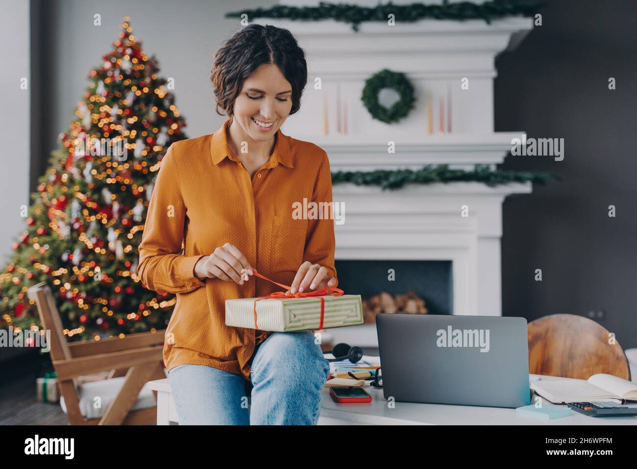 Smiling glad italian female tying red ribbon on Christmas present while sitting at home office Stock Photo