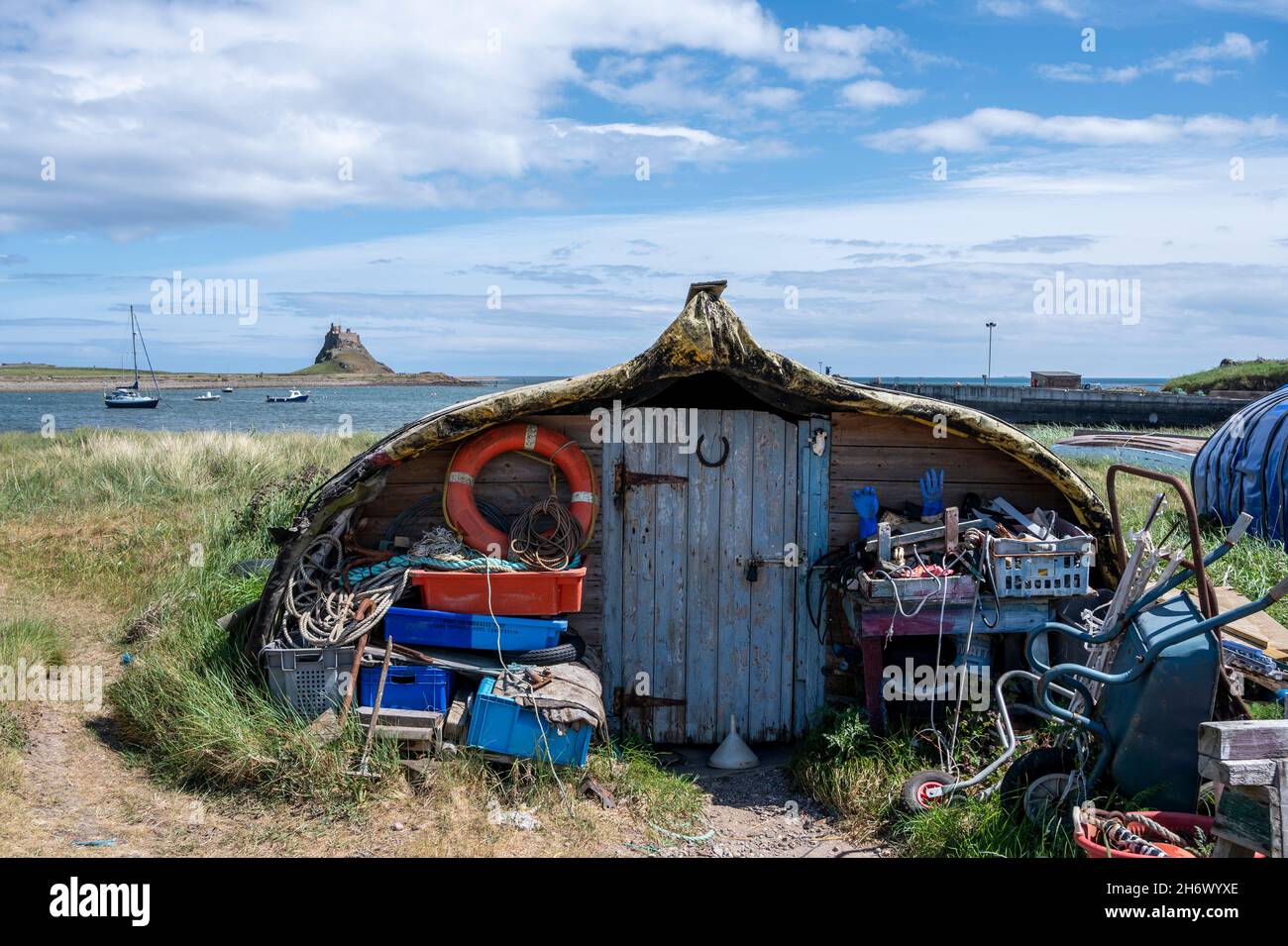 Attractive 'boat sheds' made from repurposed upturned wooden herring boats, Lindisfarne Harbour, Holy Island. Lindisfarne Castle is in the background. Stock Photo