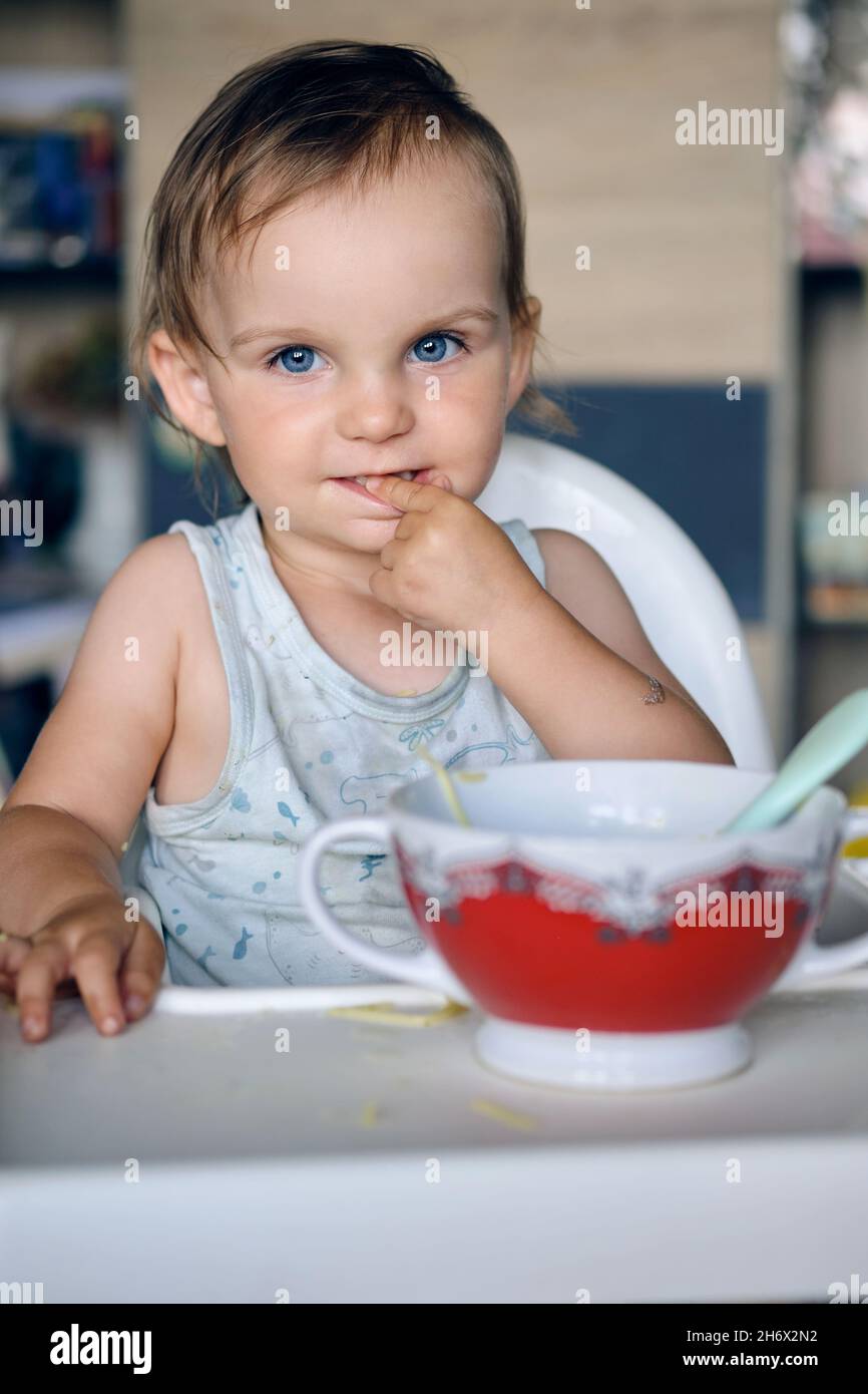 Portrait of a lovely adorable blonde little girl sitting over a bowl of soup and looking curiously at the camera Stock Photo