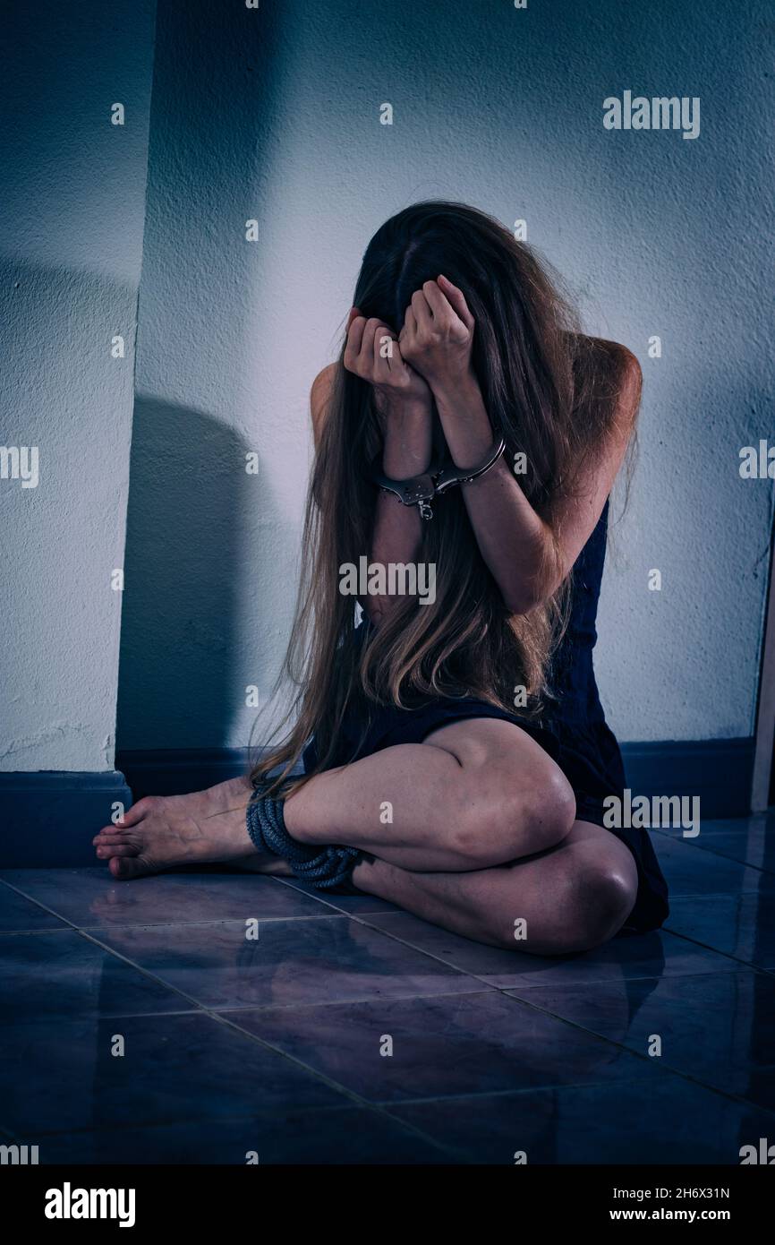 A young woman suffers from violence, sits on the floor, covers her face with hands in handcuffs. Her hands are veins and scars, her legs are tied with Stock Photo