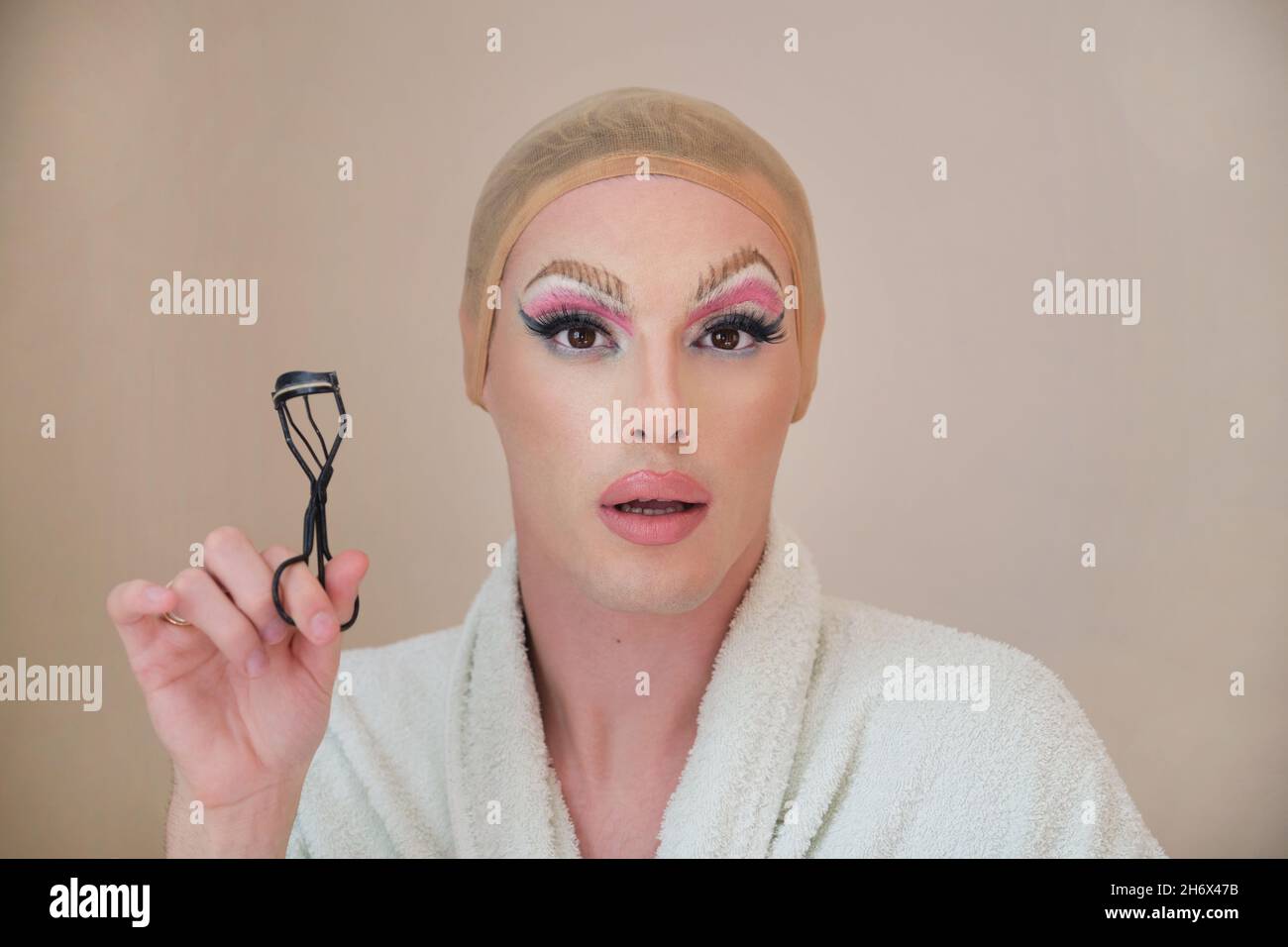 Drag queen person with a eyelash curler and wearing bathrobe looking at camera. Stock Photo