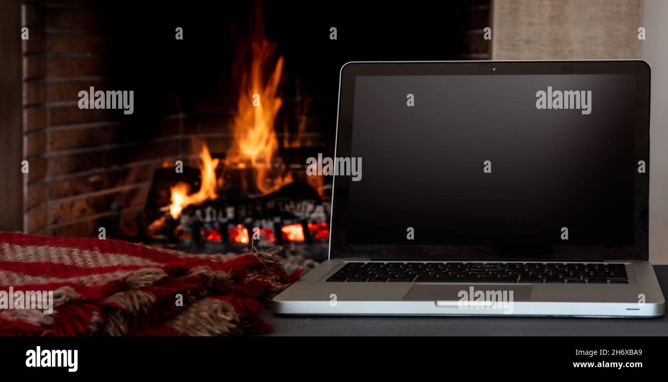Winter home office concept. Blank screen laptop on burning fireplace