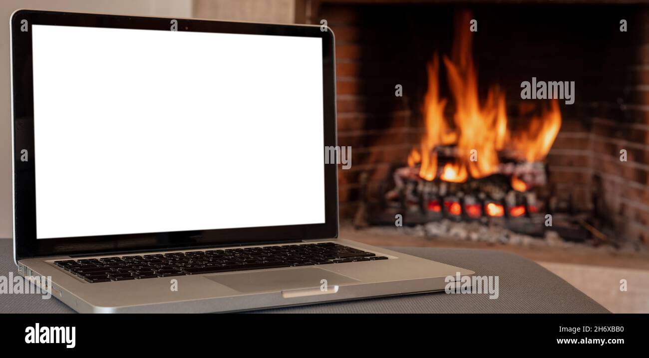 Winter home office concept. Blank screen laptop on burning fireplace
