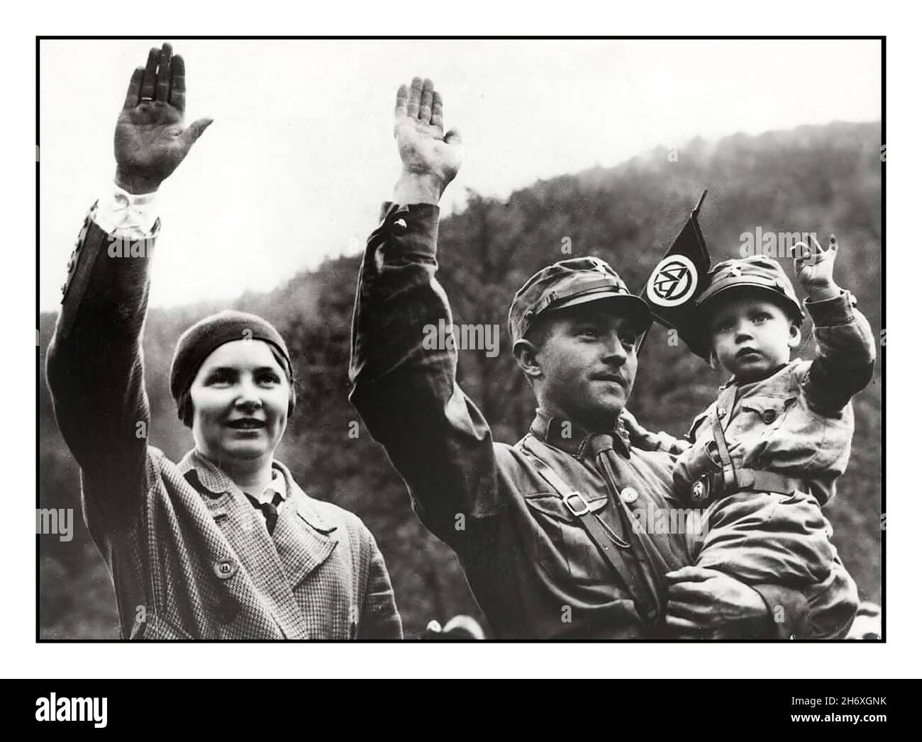 SA Sturmabteilung 1930s Archive Nazi German family of three giving the Heil Hitler salute, with the father wearing the uniform of the SA Sturmabteilung brown shirts paramilitary nazi wing. His son wearing similar infant uniform waving the flag pennant of the SA Sturmabteilung Nazi Germany. The considered ideal Aryan German Family of the 1930s Stock Photo
