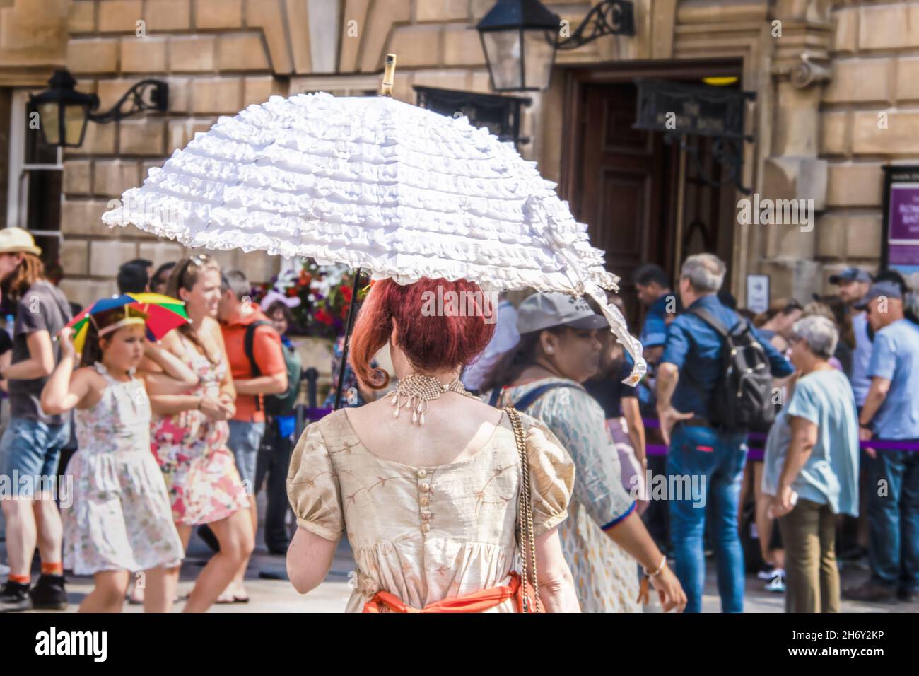 2019_07_25 Bath UK back of woman dressed in Regency Jane Austen period clothing with white ruffled parasol walking among crowd of tourists Stock Photo
