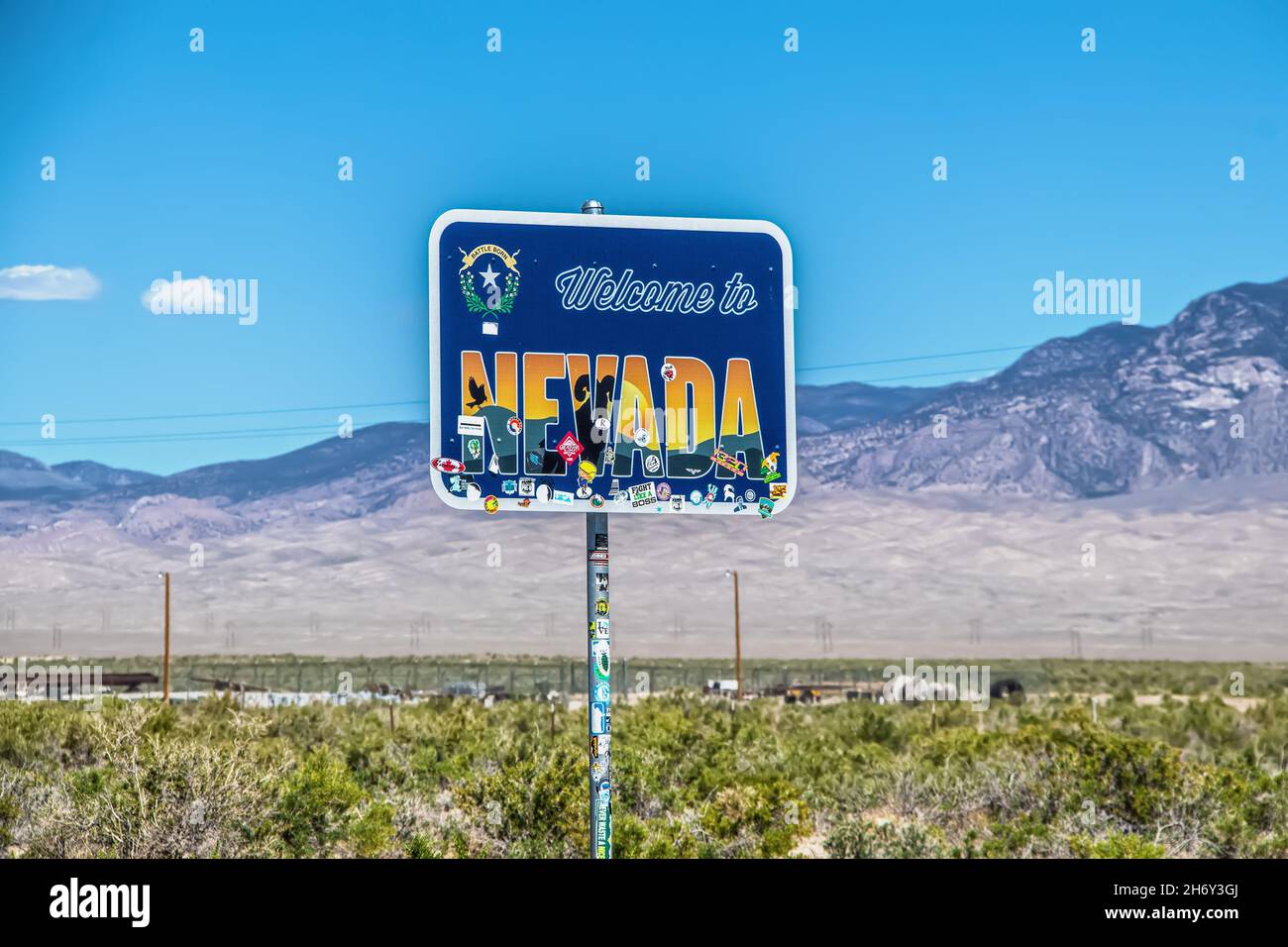 2021 06 Nevada border USA - Welcome to Nevada sign with stickers partly covering it with wire fence and desolate purple mountains blurred in backgroun Stock Photo
