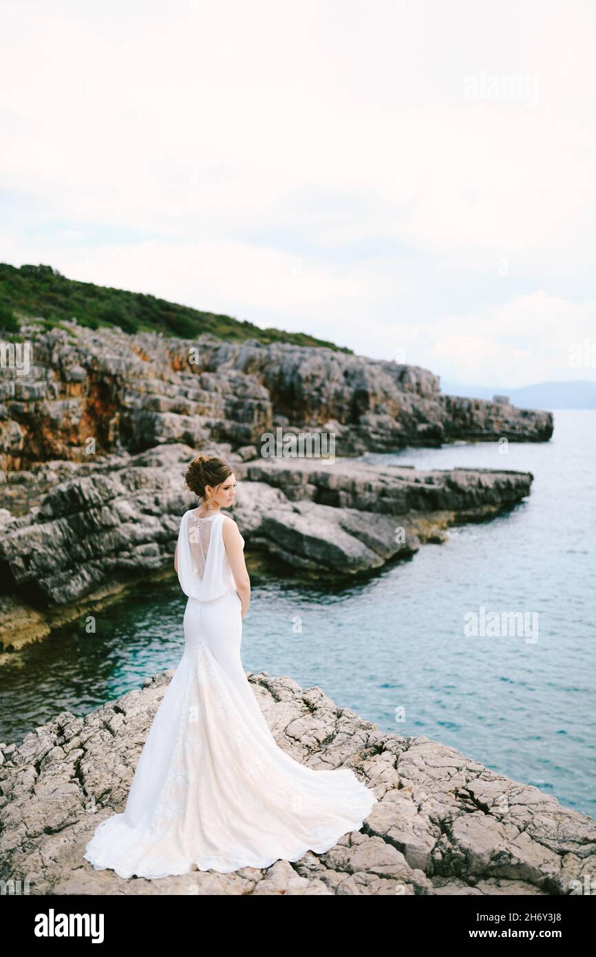 Pensive bride in a long lace white dress stands on a stone shore above ...