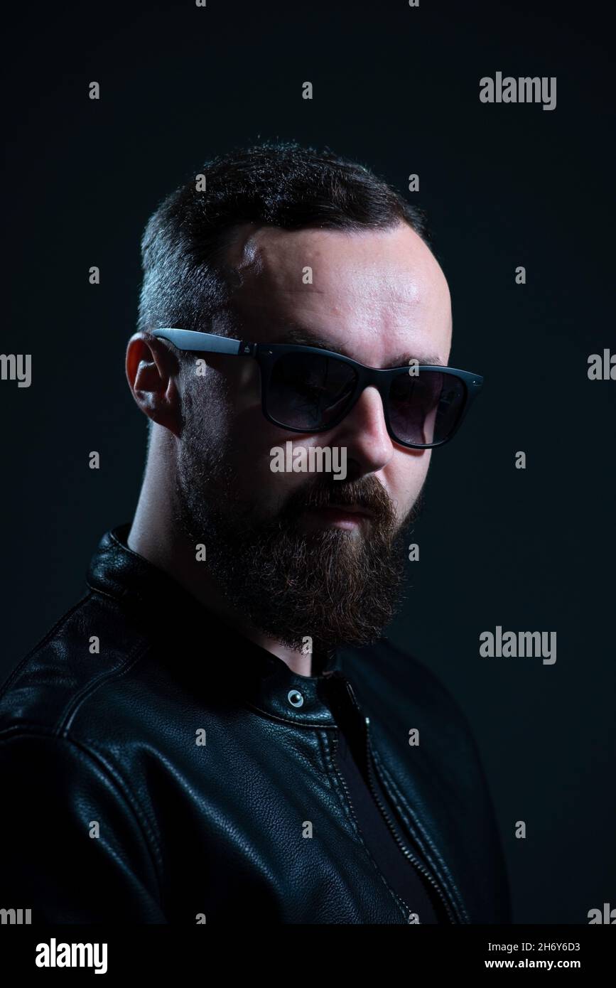 dramatic portrait of bearded millennial guy in leather jacket on dark background. Stock Photo