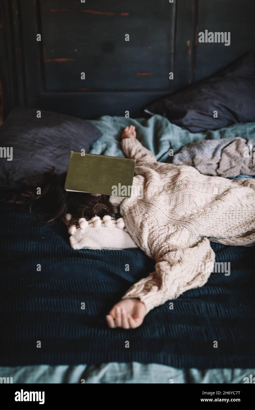 A young brunette woman in a cozy knitted sweater lying on the bed with a book on her face. Home aesthetics. Winter cozy concept Stock Photo