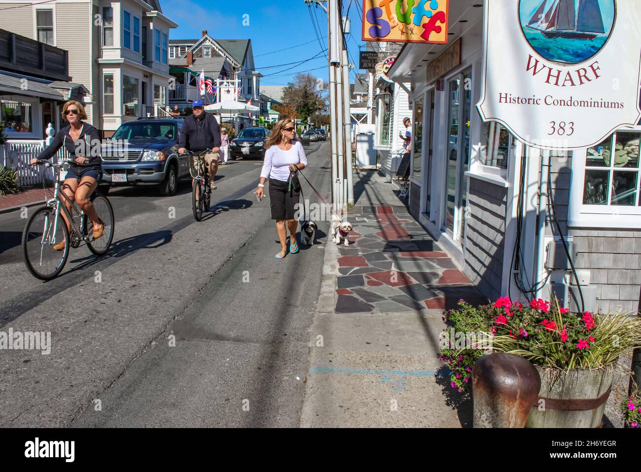 Tourists walking and bicycling down the main street looking into shops with cars parked along the street and cute dogs on leashes in Provincetown MA U Stock Photo