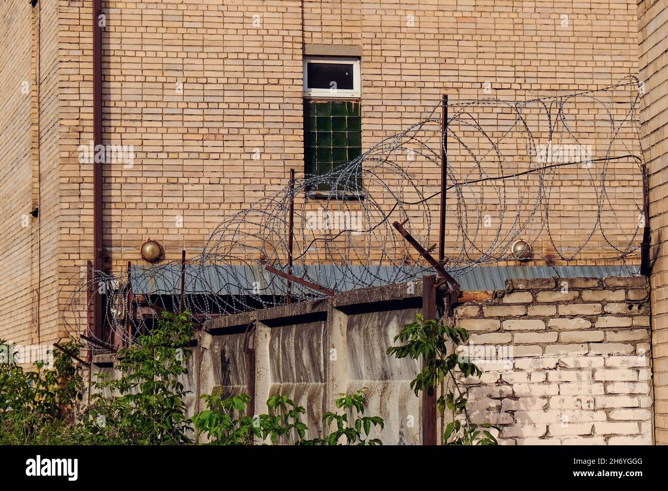 Brick wall of abandoned prison with barred window and fence with rusty barbed wire. Stock Photo