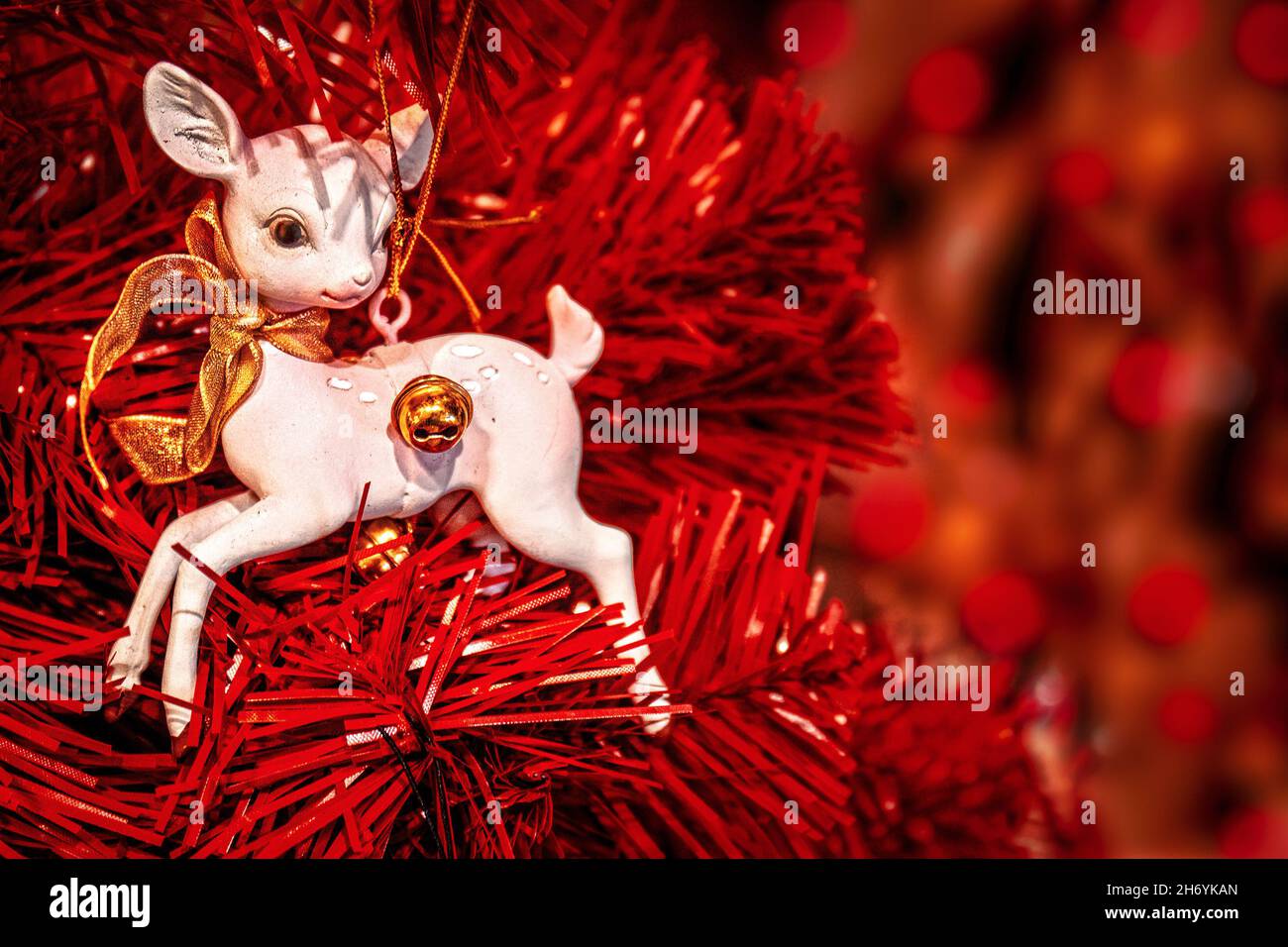 White deer ornament with gold bow on red Christmas tree to one edge of frame with bokeh background - Room for Copy. Stock Photo