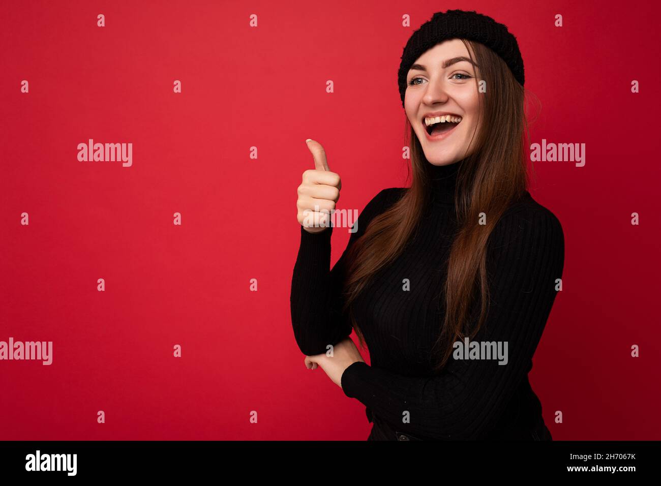 Photo of adult positive happy beautiful brunette female person with sincere emotions wearing black roll-neck sweater and black hat isolated over red Stock Photo