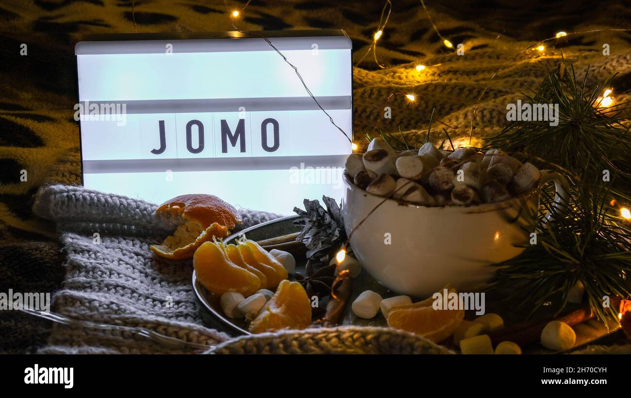 Lightbox with text JOMO Joy of missing out Cup with hot winter cacao and marshmallows tangerines spruce branch on bed. Christmas lights. Pine cones Stock Photo