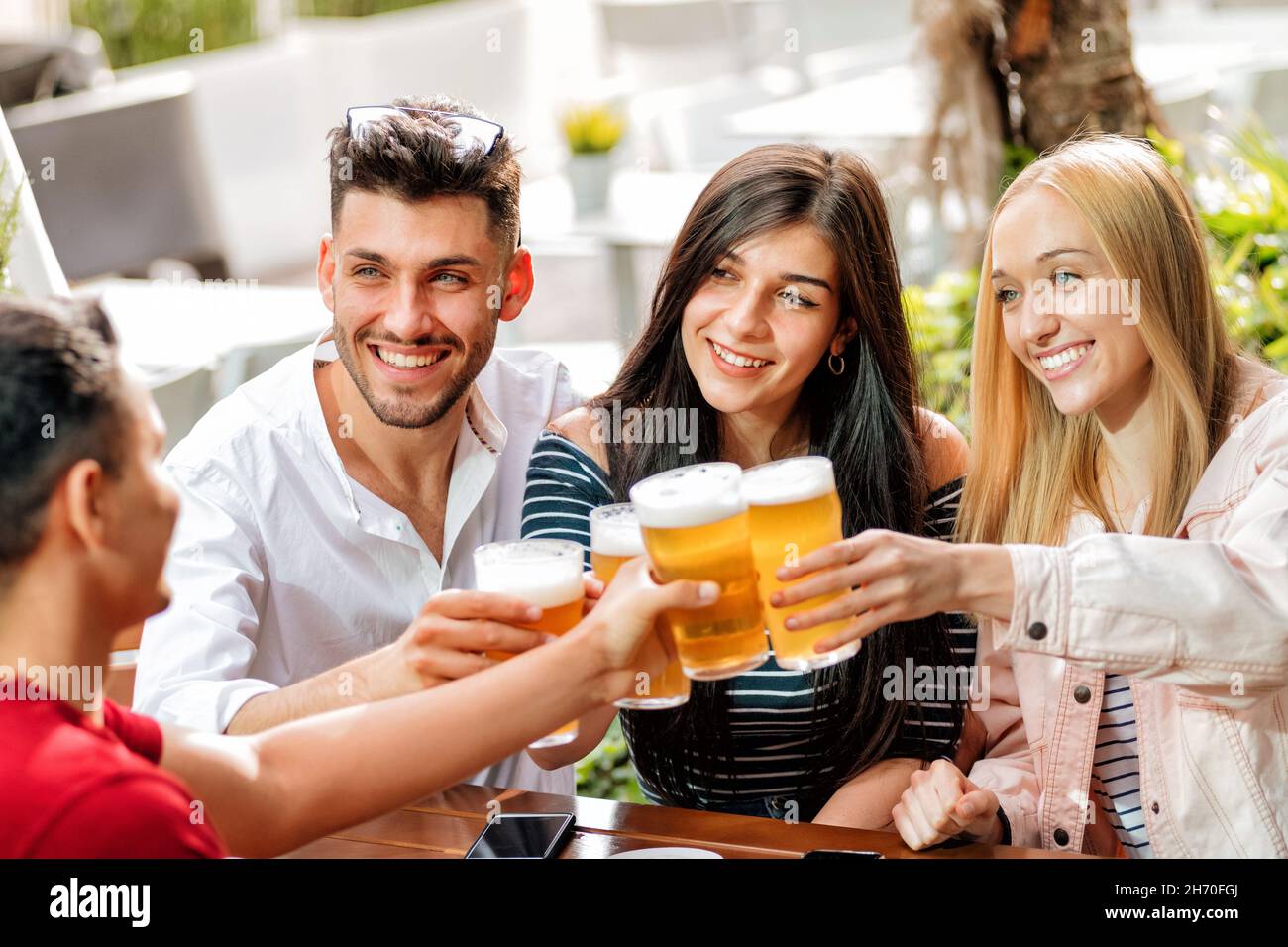 Group of positive friends in casual clothes sitting at table and clinking glasses of beers while celebrating birthday in bar in daytime Stock Photo