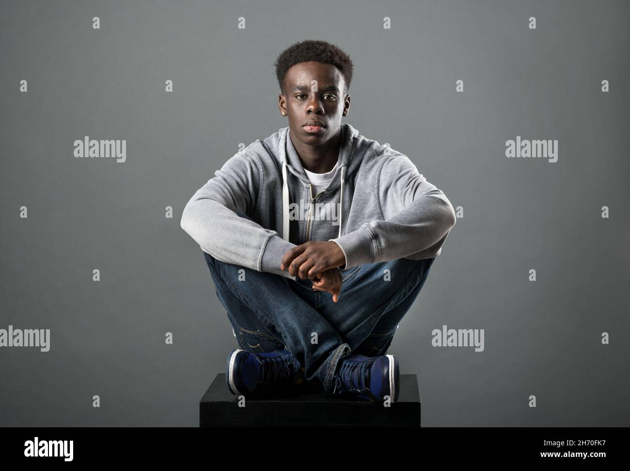 Young Black man in grey hoodie and jeans posing cross-legged in a studio looking at the camera with a serious deadpan expression Stock Photo