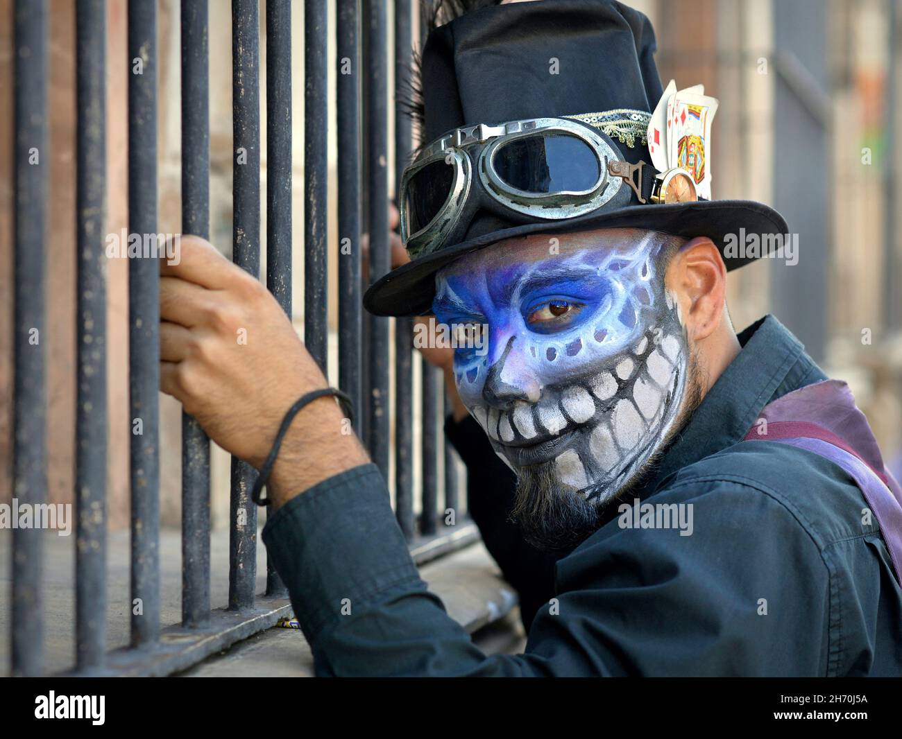 Costumed young Mexican Yucatecan man with spooky blue face painting holds onto strong burglar bars on the Day of the Dead (Día de los Muertos). Stock Photo