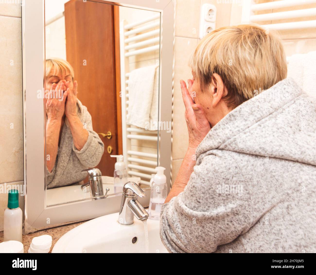 Senior woman in bathrobe touching her soft face skin, looking in mirror at home Stock Photo