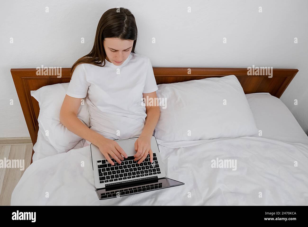 Top view of cute caucasian young woman sitting on comfy white bed at home typing on keyboard uses laptop for chatting. Millennial brunette lady workin Stock Photo