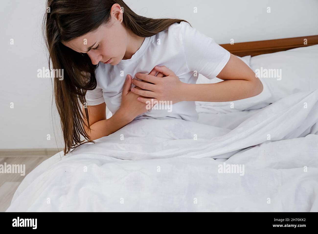 Upset tired young brunette female feeling pain ache touching chest having heart attack, sad unhappy worried girl suffers from heartache sit on bed Stock Photo
