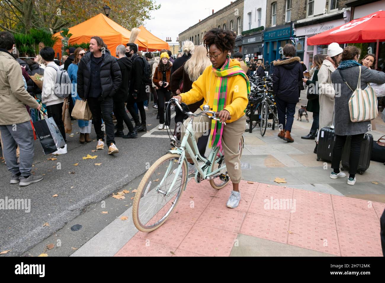 Columbia Road Flower Market young woman yellow top scarf street fashion on bike by plant stalls Sunday in November 2021 East London UK KATHY DEWITT Stock Photo