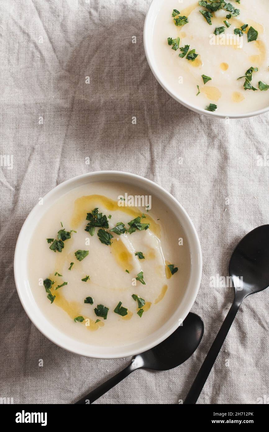 Two bowls of celeriac soup topped with olive oil and chopped parsley served with black soup spoons. Stock Photo