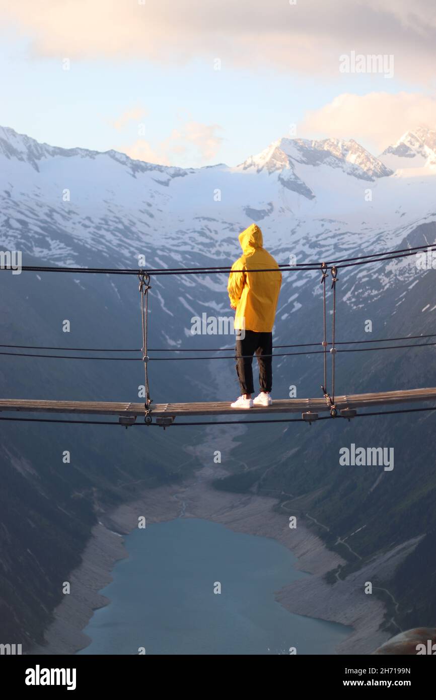 Man stands alone with a yellow rain jacket on a suspension bridge. Olperer Stock Photo