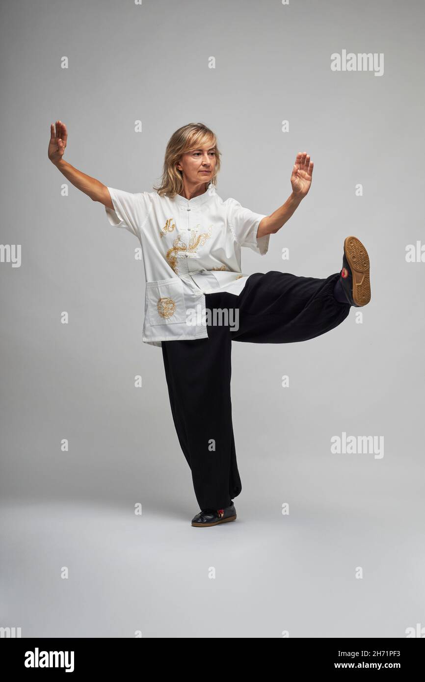 mature blonde woman practicing chi kung and tai chi on a white background she wears a traditional white chinese tai chi jacket black trousers and bl 2H71PF3