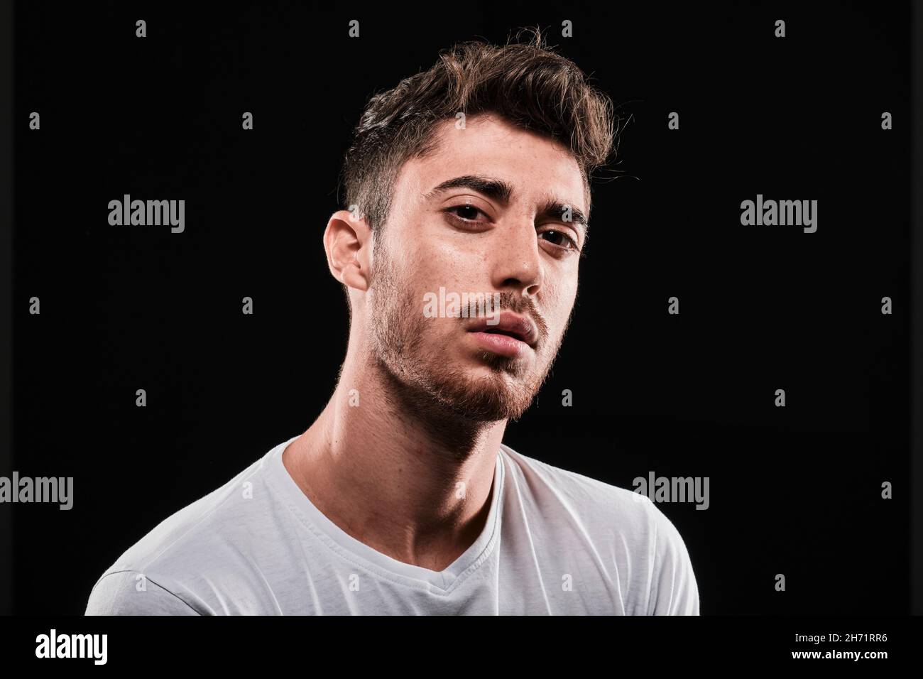 Young man close up portrait on a black background. He is wearing a basic white tshirt and looking at camera. Stock Photo
