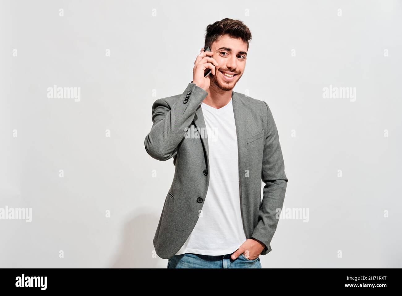 Happy young man wearing jeans, a white tshirt and a grey blazer talking with someone on his mobile phone Stock Photo