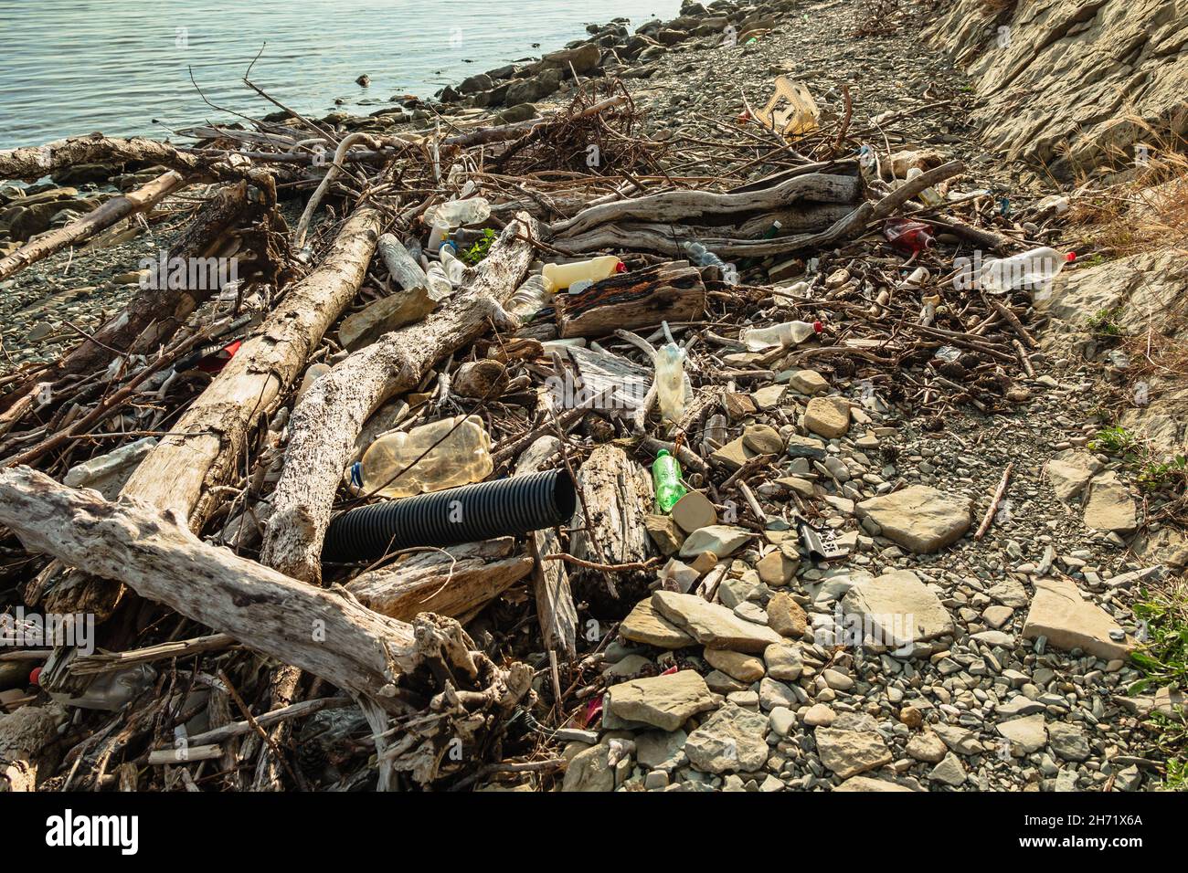 Logs, branches, plastic bottles and other debris are lying on the seashore. The stone beach is littered with garbage. The concept of ecology Stock Photo