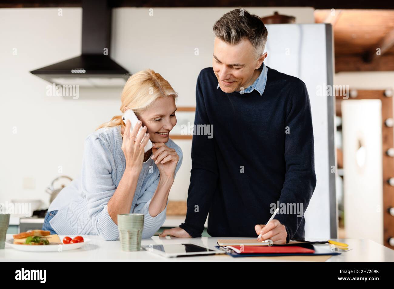 Smiling mid aged couple using cellphone and making notes in planner in kitchen Stock Photo