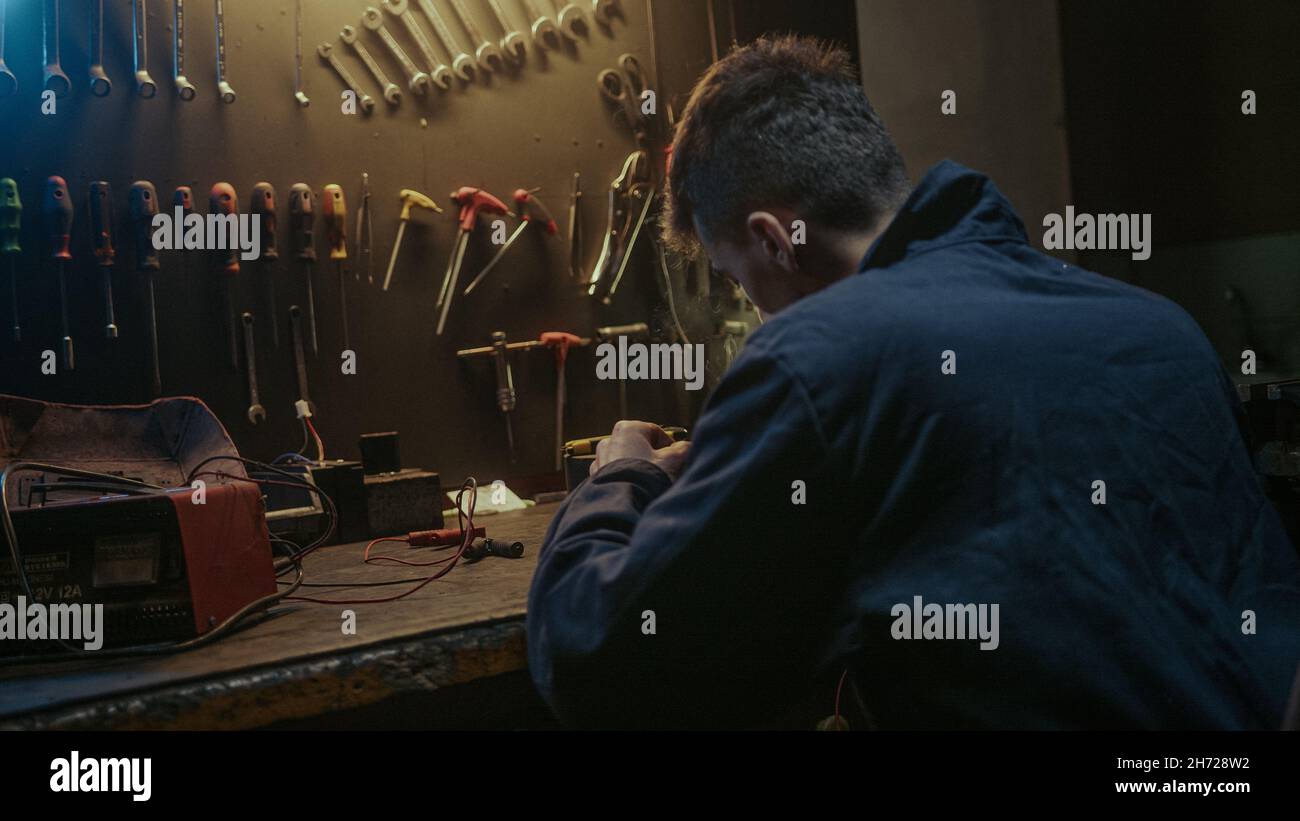 Young workshop owner man with workwear, sitting on chair and solder on workbench Stock Photo