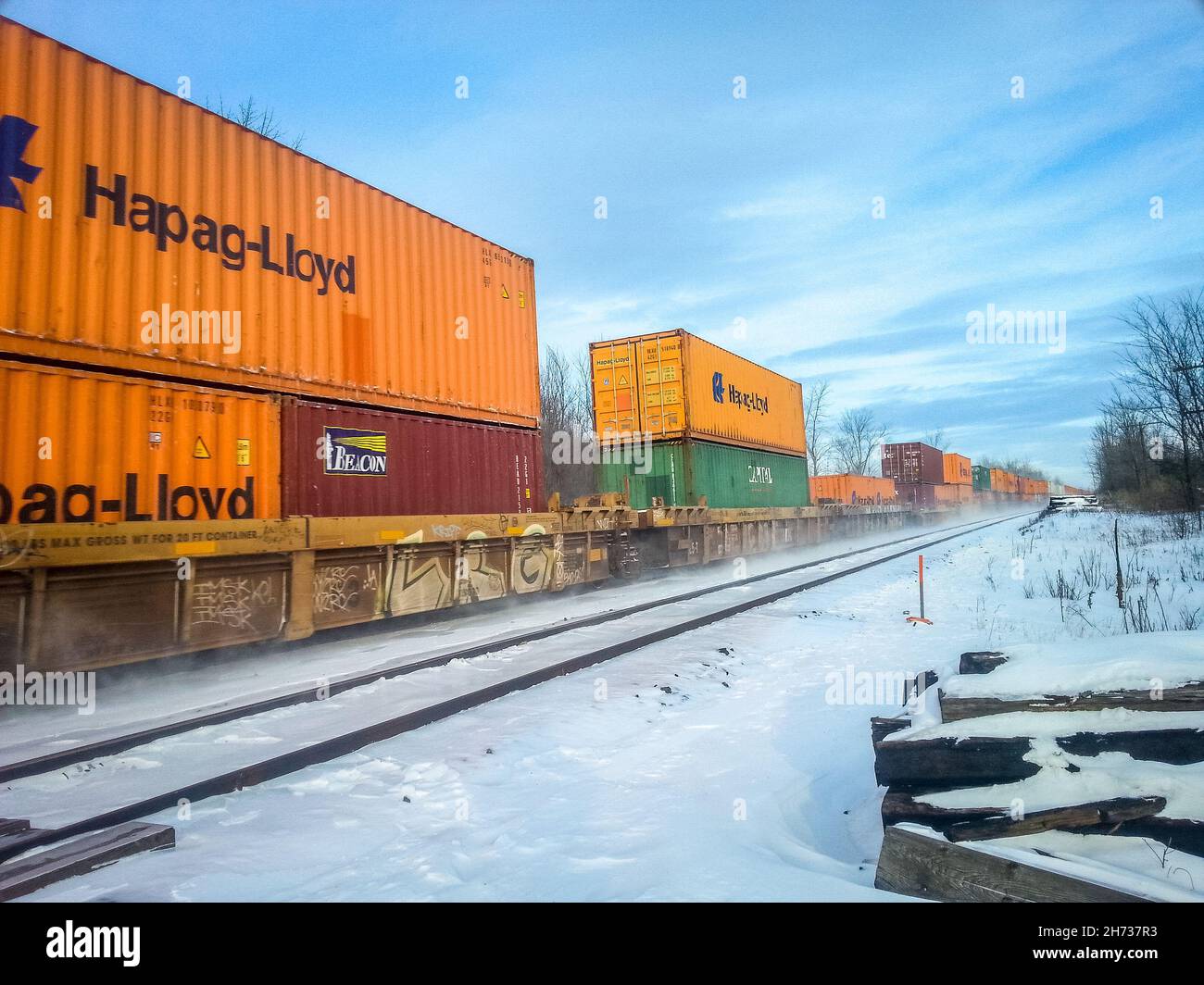 Angled view of railcars on the left side of photo, with orange, red and green box cars travelling on the train track on a sunny, snowy, winter day. Stock Photo