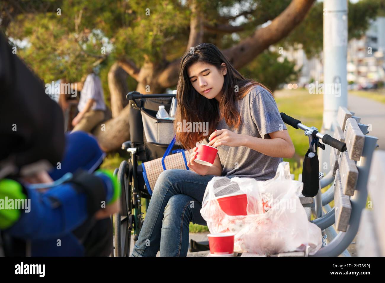 Biracial teen girl sitting outdoors on park bench eating food from red containers, outdoor dining Stock Photo