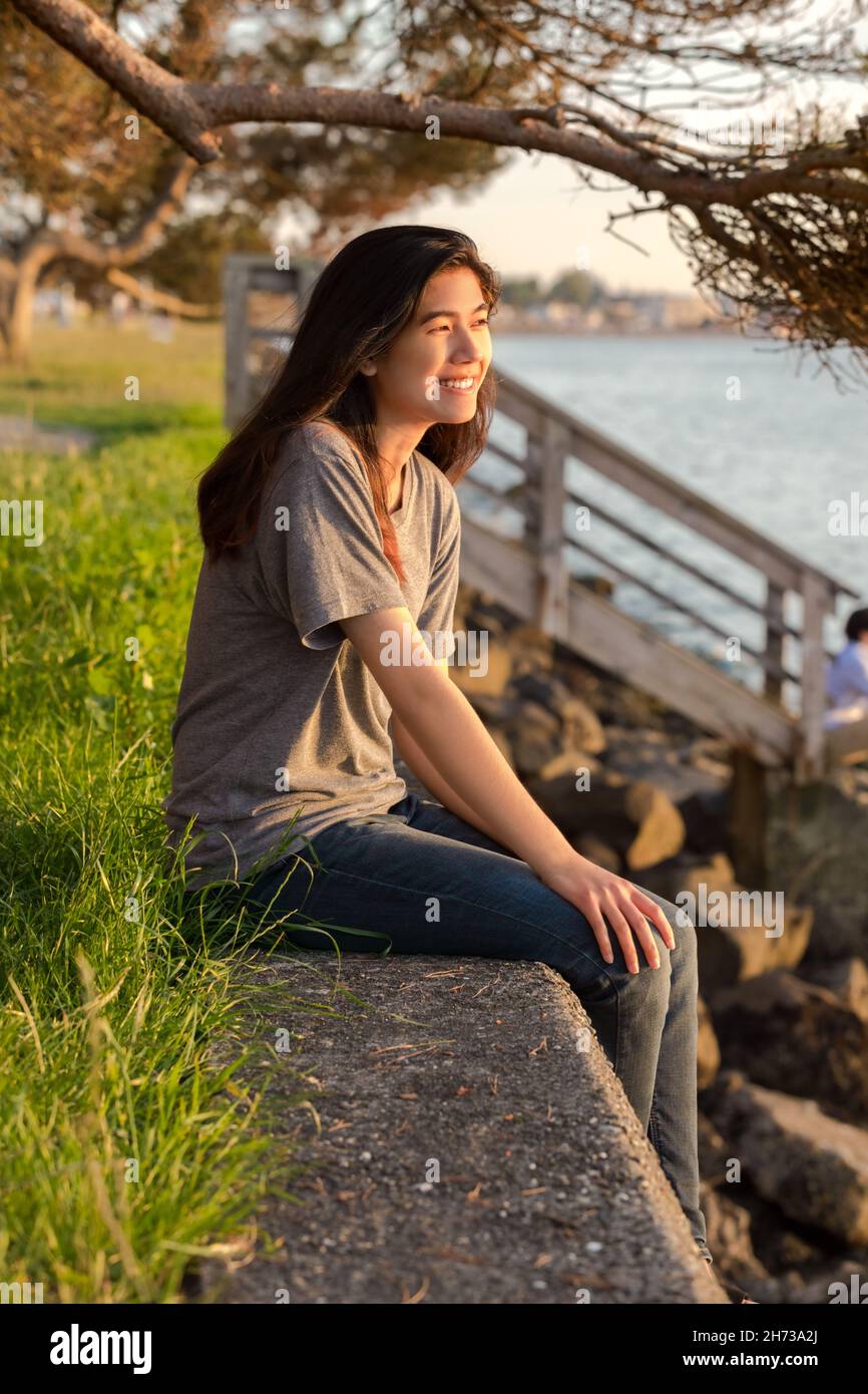 Biracial Asian Caucasian teen girl sitting on stone wall by lake at sunset Stock Photo