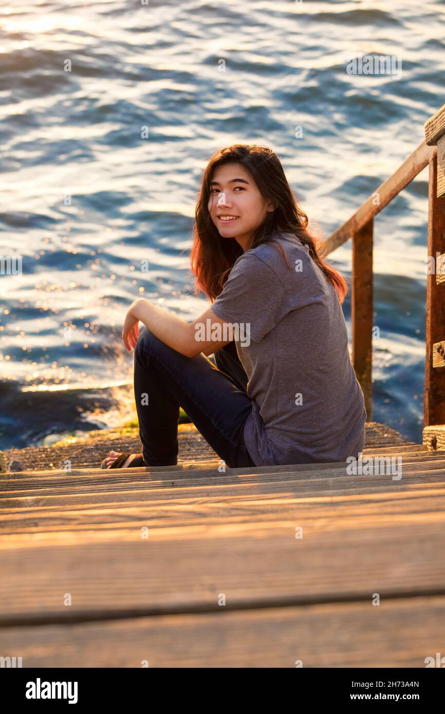 Biracial Asian teen girl relaxing on stone steps leading down to water Stock Photo