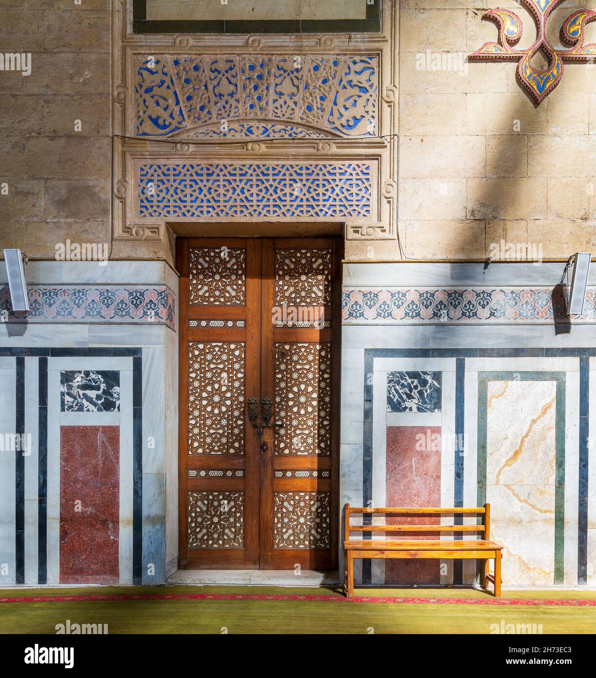 Ornamental wooden door, marble mosaic decorated walls, wooden bench, and floor lit by sunlight beams, in public historic Al Rifai Mosque, Cairo, Egypt Stock Photo