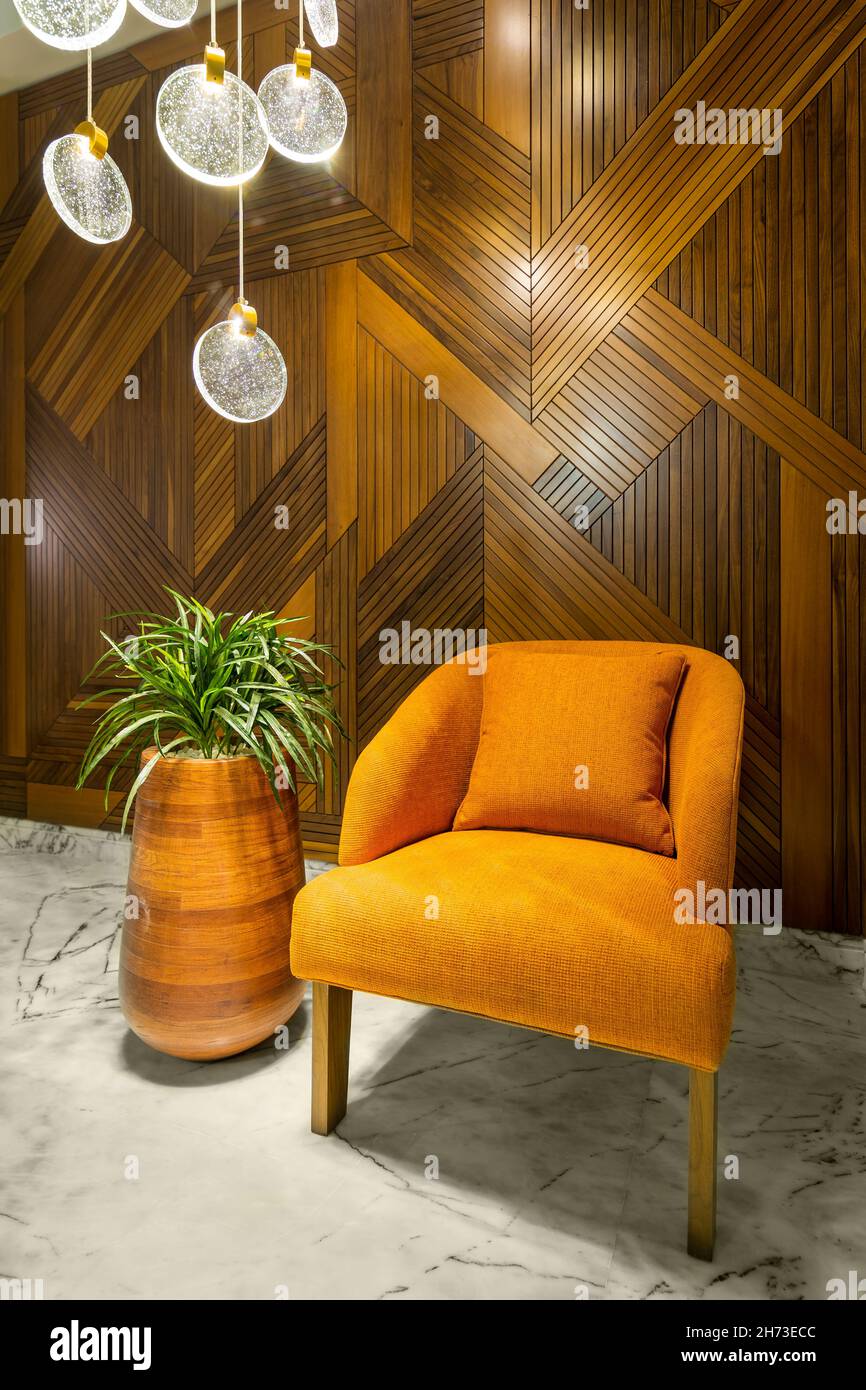 Modern orange armchair, tall wooden planter with green bushes, and contemporary glass chandelier, in a hall with decorated wood cladding wall, and white marble floor Stock Photo
