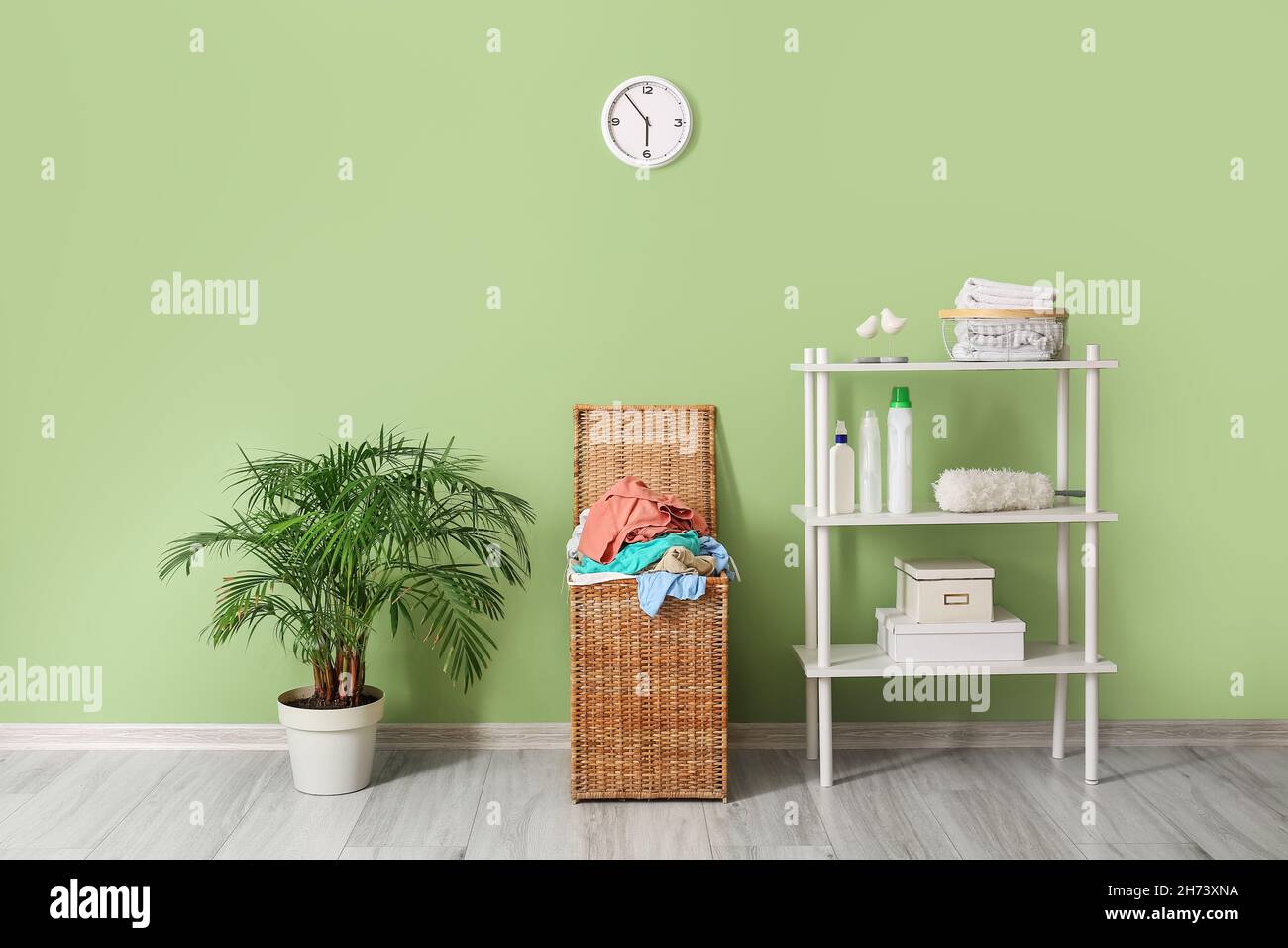 Wicker basket with laundry, shelving unit and houseplant near green wall Stock Photo