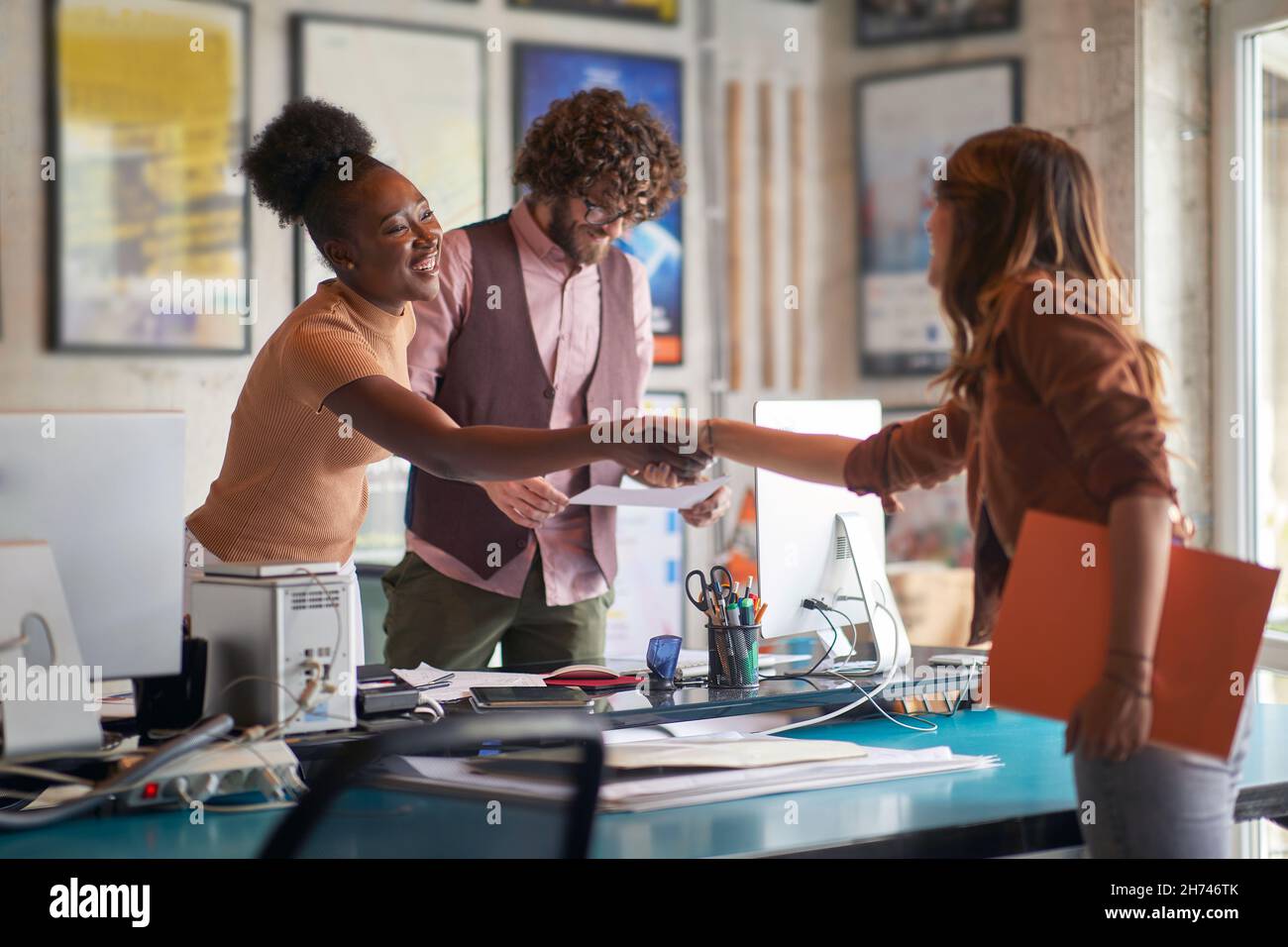 afro american and caucasian females shaking hands over the desk in office with beardy male holding and reading document Stock Photo