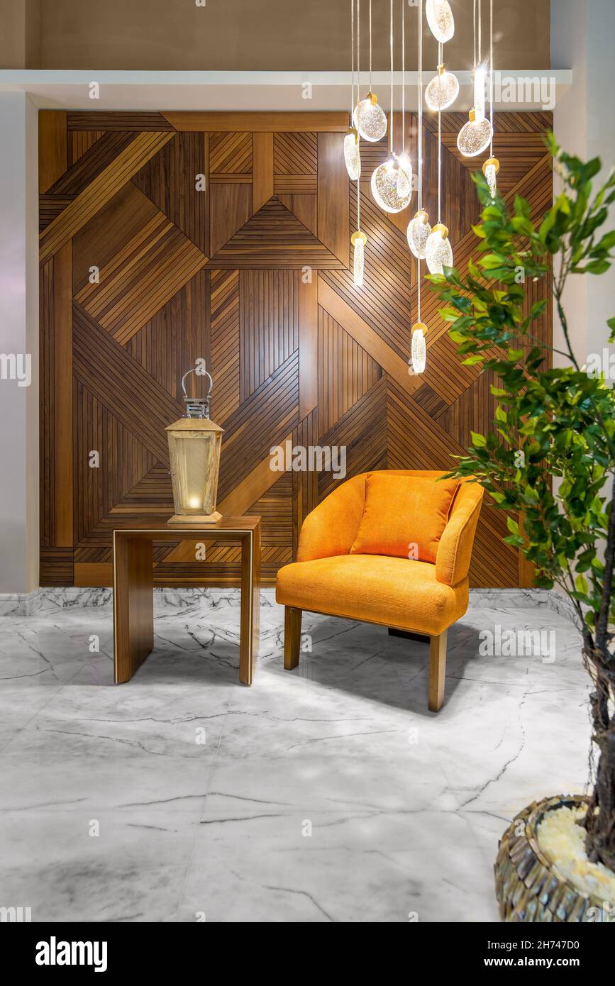 Modern orange armchair, illuminated wooden lantern on small wooden table, and contemporary tall glass chandelier, in a hall with decorated wood cladding wall, and white marble floor Stock Photo