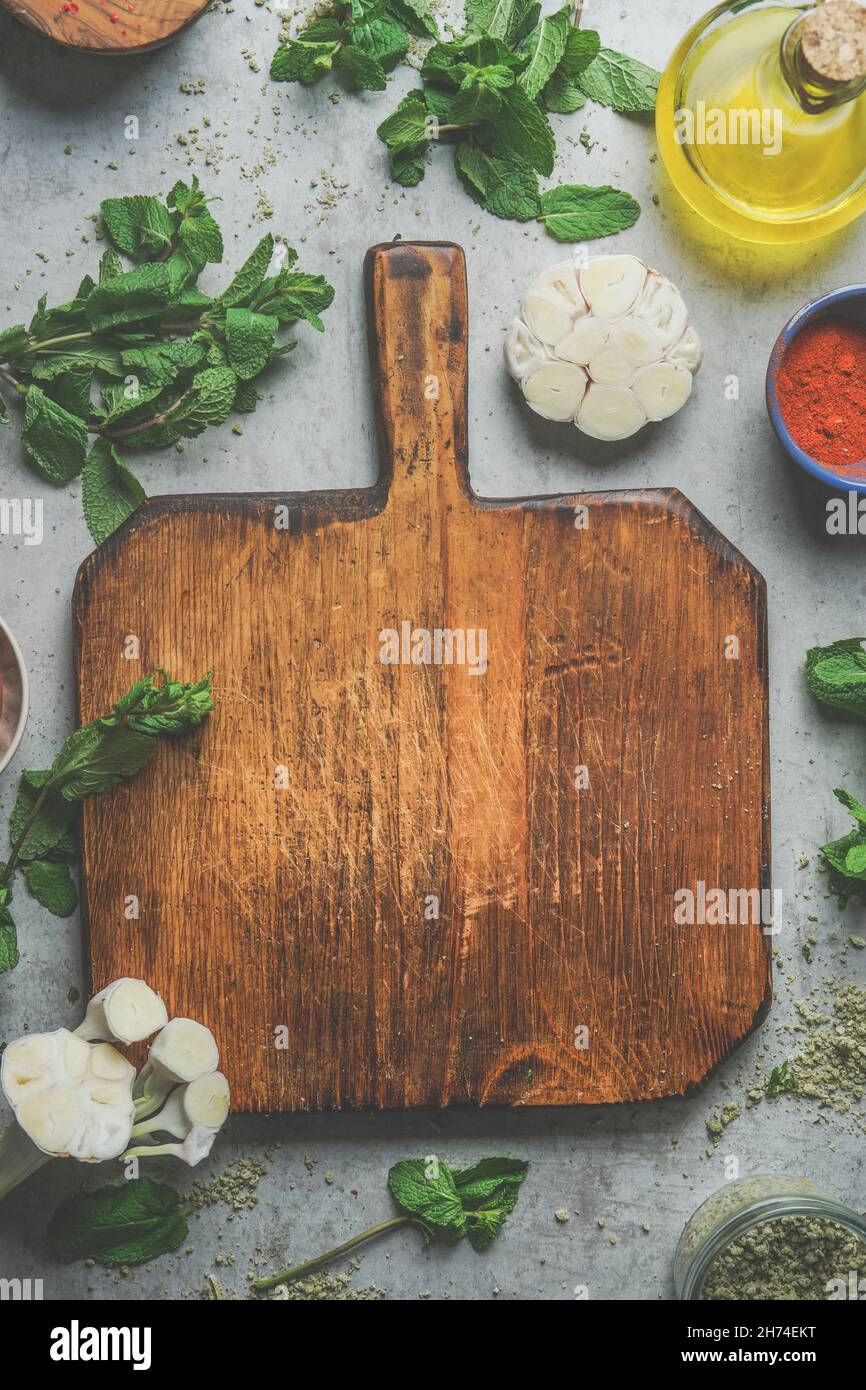 Food background with empty wooden cutting board, herbs, garlic, olive oil, spices on grey concrete kitchen table. Cooking with fresh, flavorful ingred Stock Photo