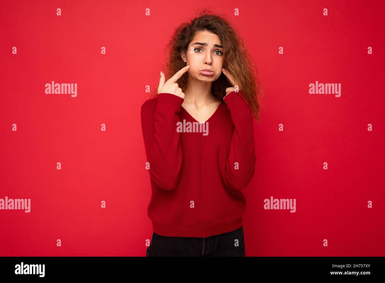 Photo of young offended touchy sad attractive brunette curly woman with sincere emotions wearing casual red pullover isolated on red background with Stock Photo