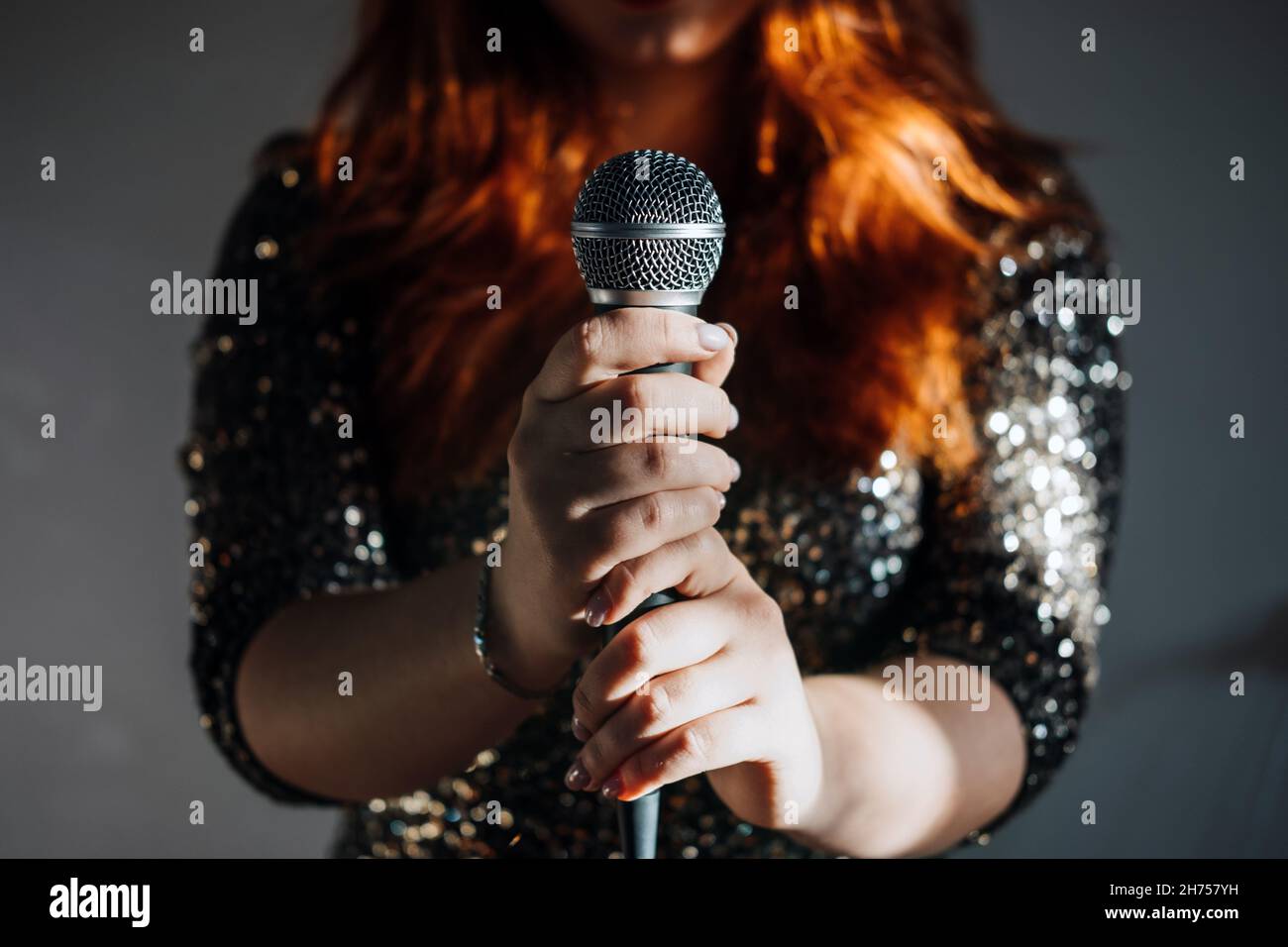 Faceless portrait of redhead woman in sparkly evening dress holding microphone on dark night background. Unrecognizable female singer with modern Stock Photo