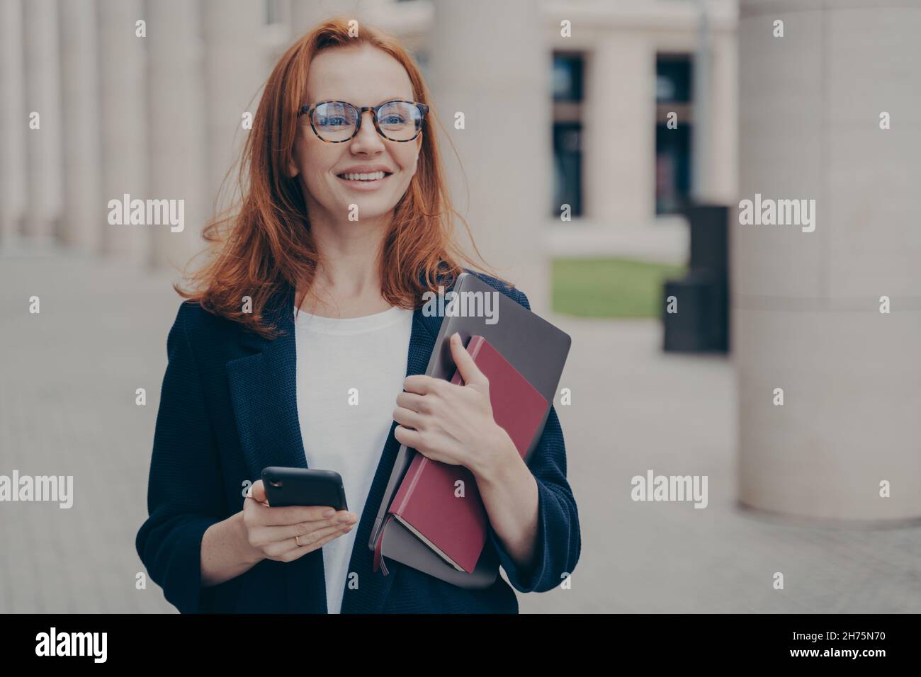 Happy red-haired business lady in official clothes standing outdoors, using mobile phone Stock Photo