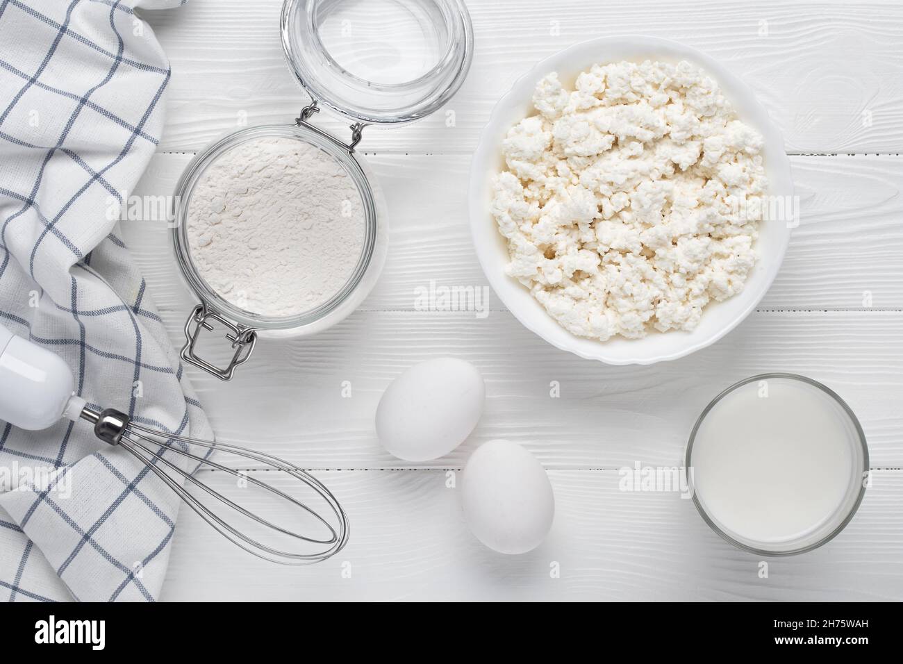 Raw curd cheese, flour, eggs, milk and whisk on a white wooden table. Cooking concept. Recipe ingredients. Baking products. View from above. Flat lay Stock Photo