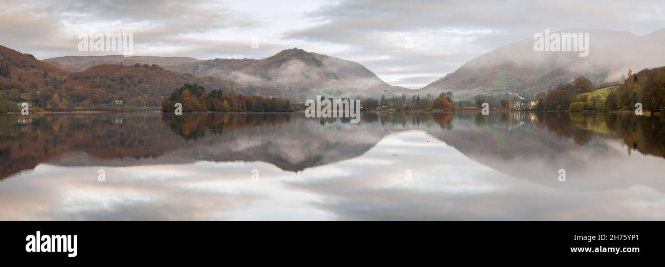 Grasmere village and the surrounding fells are perfectly reflected in the still waters of the lake on a calm autumn morning as the mist slowly clears. Stock Photo